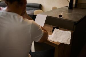 arrière vue de une homme en cours d'analyse le les dépenses et frais pour payant factures, séance à bureau à Accueil avec les documents dans mains photo