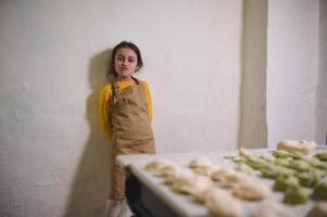 malicieux peu futur chef femme au foyer dans beige cuisine tablier, permanent contre blanc mur arrière-plan, souriant et fabrication visages à caméra. sur le premier plan une table avec sculpté Dumplings varenniki photo