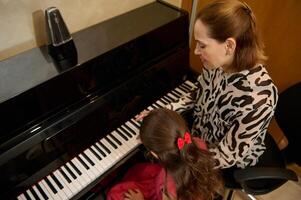 arrière vue de au dessus de une peu enfant fille et la musique prof en jouant ensemble le grandiose piano pendant individuel la musique leçon à Accueil photo