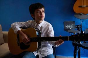 sérieux adolescente garçon performant du son mélodie, en chantant chanson tandis que en jouant acoustique guitare dans moderne Accueil musical studio photo