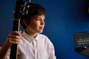 adolescent garçon musicien guitariste dans blanc chemise, rêveusement à la recherche de côté, posant avec acoustique guitare bleu mur Contexte photo