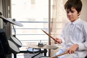 authentique portrait de adolescent garçon en jouant musical percussion instrument avec des bâtons, séance par électrique tambour ensemble contre fenêtre Contexte. magnifique éclairage sur étape photo