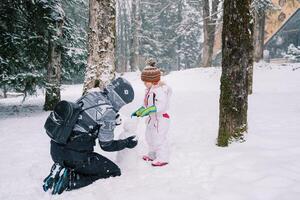 peu fille aide sa mère sculpter une bonhomme de neige dans une neigeux forêt photo