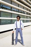 travailleur masculin noir debout à côté d'un immeuble de bureaux avec une planche à roulettes. photo