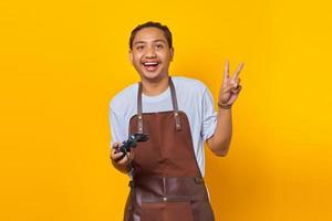portrait de joyeux beau jeune homme asiatique portant un tablier tenant un contrôleur de jeu et montrant le numéro deux avec le doigt photo