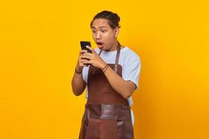 portrait d'un beau barista qui est choqué et surpris de voir une promotion sur un smartphone sur fond jaune photo