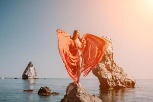 femme Voyage mer. Jeune content femme dans une longue rouge robe posant sur une plage près le mer sur Contexte de volcanique rochers, comme dans Islande, partage Voyage aventure périple photo