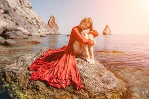 femme Voyage mer. content touristique dans rouge robe prendre plaisir prise image en plein air pour souvenirs. femme voyageur posant sur le Roche à mer baie entouré par volcanique montagnes, partage Voyage aventure périple photo