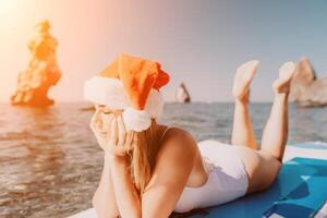 femme mer souper. proche en haut portrait de content Jeune caucasien femme avec longue cheveux dans Père Noël chapeau à la recherche à caméra et souriant. mignonne femme portrait dans une blanc bikini posant sur souper planche dans le mer photo