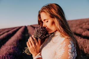 femme lavande champ. content insouciant femme dans une blanc robe en marchant dans une lavande champ et odeur une lavande bouquet sur le coucher du soleil. idéal pour chaud et inspirant concepts dans esprit d'aventure et Voyage. photo