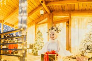 content souriant femme dans une blanc robe en portant une cadeau boîte dans de face de une Noël arbre. photo