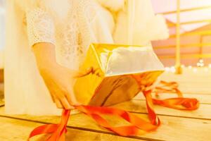 une femme dans une blanc robe est en portant une or boîte avec une rouge ruban. elle est portant une couronne sur sa diriger. le femme apparaît à être ouverture le cadeau boîte. photo