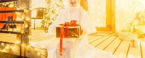 content souriant femme dans une blanc robe en portant une cadeau boîte dans de face de une Noël arbre. photo