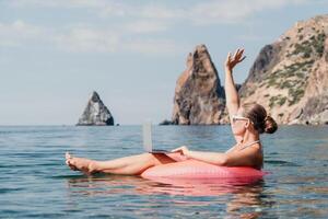 femme pigiste travaux sur portable nager dans mer sur rose gonflable anneau. content touristique dans des lunettes de soleil flottant sur gonflable Donut et travail sur portable ordinateur dans calme océan. éloigné travail nulle part photo