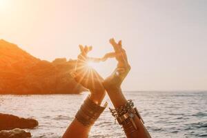 femme mer yoga. content femme dans une blanc maillot de bain et style bohème bracelets fabrication une cœur forme avec sa mains tandis que pratiquant yoga sur une tapis par mer à le coucher du soleil. en bonne santé mode de vie, harmonie et méditation photo