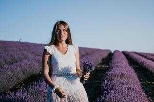 femme lavande champ. content insouciant femme dans une blanc robe en marchant dans une lavande champ et odeur une lavande bouquet sur le coucher du soleil. idéal pour chaud et inspirant concepts dans esprit d'aventure et Voyage. photo