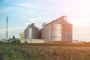 grain silos sur une vert champ Contexte avec chaud le coucher du soleil lumière. grain ascenseur. métal grain ascenseur dans agricole zone. agriculture espace de rangement pour récolte. aérien vue de agricole usine. personne. photo