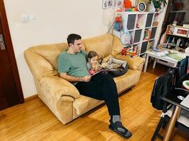 papa lit une livre à une peu fille séance sur le canapé suivant à une mensonge chat photo