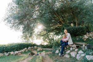 souriant mère avec une peu fille dans sa bras est assis sur une parc banc suivant à une tigré chat photo