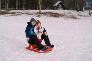 souriant papa avec peu fille sont séance sur une traîneau sur une neigeux plaine photo