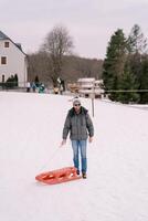 souriant homme dans des lunettes de soleil avec traîneau des stands sur une couvert de neige colline près une corde ascenseur photo