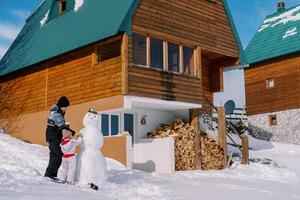 maman et peu fille décorer une bonhomme de neige près une tas de bois de une en bois chalet dans le neige photo
