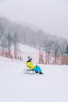 peu garçon dans une coloré ski costume monte une traîneau vers le bas le pente de une neigeux Montagne. côté vue photo