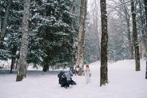 peu fille des stands dans une neigeux forêt et regards à sa maman fabrication une bonhomme de neige photo