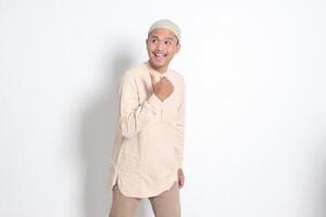 portrait de sous le choc asiatique musulman homme dans koko chemise avec calotte montrant produit et montrer du doigt avec pouce en haut à le côté. La publicité concept. isolé image sur blanc Contexte photo
