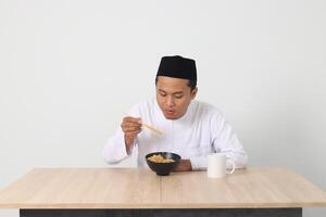 portrait de attrayant asiatique musulman homme en mangeant de bon goût instant nouilles avec baguettes servi sur bol. iftar et pré Aube repas concept. isolé image sur blanc Contexte photo