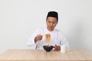 portrait de attrayant asiatique musulman homme en mangeant de bon goût instant nouilles avec baguettes servi sur bol. iftar et pré Aube repas concept. isolé image sur blanc Contexte photo