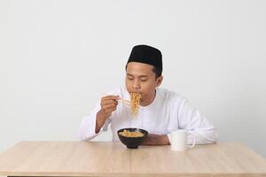 portrait de attrayant asiatique musulman homme en mangeant de bon goût instant nouilles avec baguettes servi sur bol. iftar et pré Aube repas concept. isolé image sur blanc Contexte photo