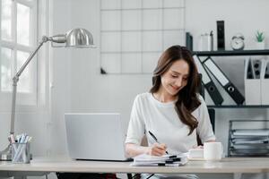 femme d'affaires en utilisant calculatrice et l'écriture Remarques suivant à sa portable dans une brillant Accueil bureau, entouré par organisé Bureau Provisions et documents. photo