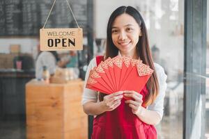 content asiatique femme entrepreneur en portant rouge enveloppes, prêt à célébrer, permanent dans de face de une accueillant ouvert signe à sa café. photo