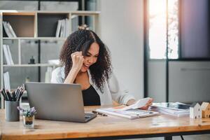 joyeux femme d'affaires est Profond dans pensée, en cours d'analyse les documents avec une sourire, dans une brillant espace de travail avec une portable et Bureau fournitures. photo