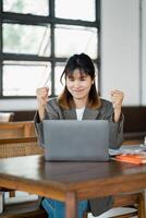 Enchanté Jeune femme soulève sa les poings dans Succès dans de face de sa portable dans une naturellement allumé, aéré espace de travail. photo