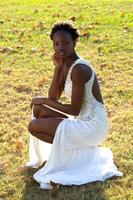 Jeune mince noir femme dans blanc robe accroupi sur herbe photo