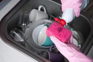 une femme dans rose caoutchouc gants verser détergent sur une cuisine éponge contre le toile de fond de sale vaisselle dans le couler. fermer. sélectif se concentrer. photo