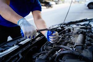 une homme est travail sur une voiture moteur. il est portant gants et une bleu chemise. le voiture est garé sur le côté de le route photo