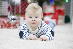 marrant aux yeux bleus enfant contre le Contexte de une floue Noël intérieur.le enfant mensonges sur le tapis et regards à le caméra. photo