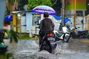 une père équitation une moto par les eaux de crue pendant lourd pluie tandis que en portant un parapluie dans une logement complexe, Indonésie, 8 décembre 2023. photo