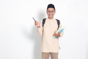 portrait de excité étudiant asiatique musulman homme dans koko chemise avec calotte porter sac à dos, en portant école livres, montrer du doigt à le côté. islamique éducation concept. isolé image sur blanc Contexte photo