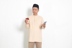 portrait de dérangé asiatique musulman homme dans koko chemise avec calotte en portant une mobile téléphone et en présentant crédit carte, trompé par en ligne prêts. isolé image sur blanc Contexte photo