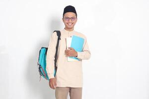 portrait de excité étudiant asiatique musulman homme dans koko chemise avec calotte porter sac à dos, tandis que en portant le sien école livres. islamique éducation concept. isolé image sur blanc Contexte photo