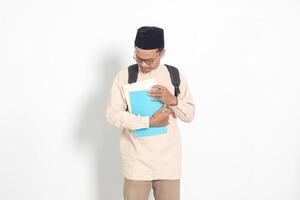 portrait de excité étudiant asiatique musulman homme dans koko chemise avec calotte porter sac à dos, tandis que en portant le sien école livres. islamique éducation concept. isolé image sur blanc Contexte photo