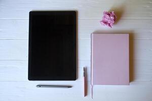 une tablette, bloc-notes, stylo et une froissé feuille de papier sur une blanc en bois bureau. photo