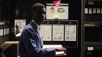 portrait de africain américain enquêteur permanent dans preuve chambre, en train de lire Cas des dossiers. dans vaguement allumé bureau, Masculin inspecteur examine suspect déclarations et regards à caméra. au ralenti, agrandir. photo