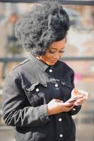 attrayant content peau foncée modèle avec afro coiffure et piercing au nez, posant en plein air contre Urbain Contexte pendant sa Matin marcher, à la recherche vers le bas avec timide sourire montrant sa blanc dents. éclater Soleil photo