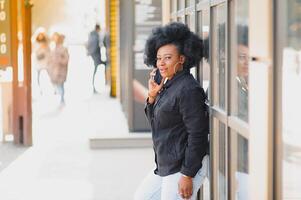 attrayant content peau foncée modèle avec afro coiffure et piercing au nez, posant en plein air contre Urbain Contexte pendant sa Matin marcher, à la recherche vers le bas avec timide sourire montrant sa blanc dents. éclater Soleil photo