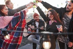 beau mec chante pendant que des amis prennent un verre. groupe de jeunes faisant la fête sur un toit avec de l'alcool et de la guitare photo